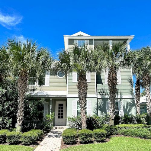 A multi-story house with palm trees and a clear blue sky in the background, surrounded by greenery and a stone pathway leading to the door.