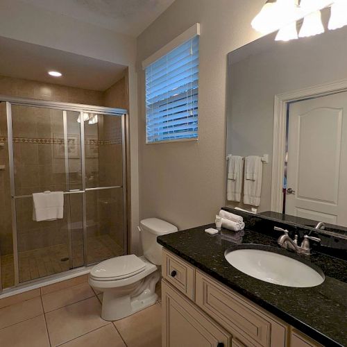 The image shows a bathroom with a shower, toilet, sink, large mirror, and towels on racks, featuring neutral colors and modern decor.