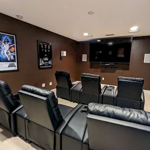 A home theater with leather recliners, a large TV, wall speakers, and movie posters on brown walls, creating a cozy viewing space.