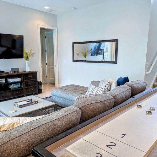 A cozy living room featuring a sectional sofa, wall-mounted TV, shuffleboard table, and stairs leading upwards, with a large mirror on the wall.