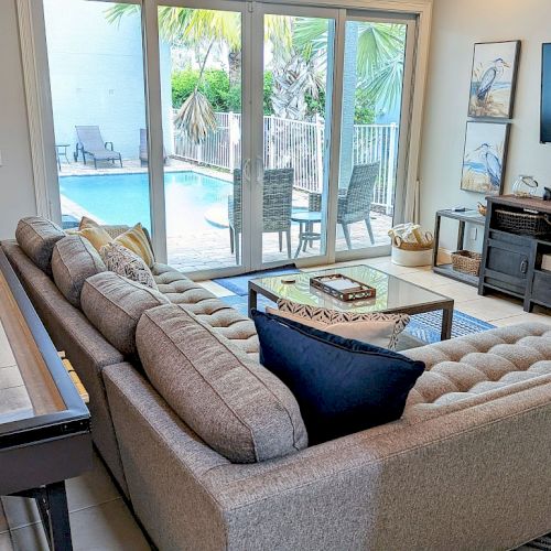 A cozy living room with a sectional sofa, shuffleboard, TV, and windows overlooking a pool area complete the inviting interior.