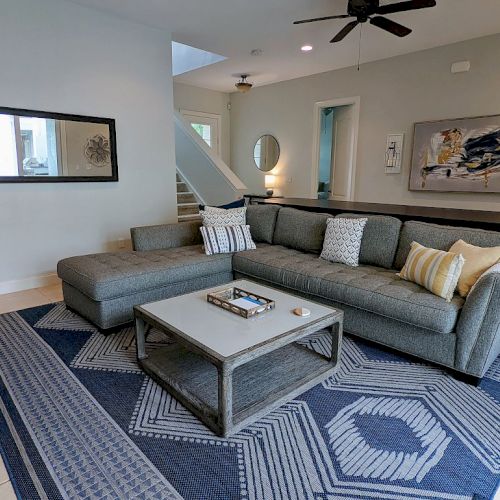 A modern living room with a gray sectional sofa, blue patterned rug, wall art, and a mirror, featuring minimalist decor accents.