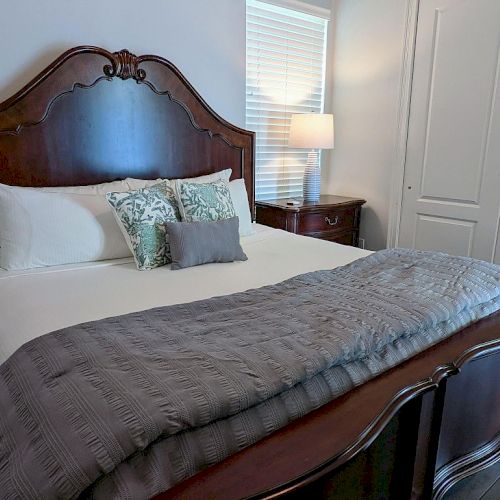 A neatly made bed with a wooden headboard, pillows, a lamp, and a side table, next to a window and closet doors.