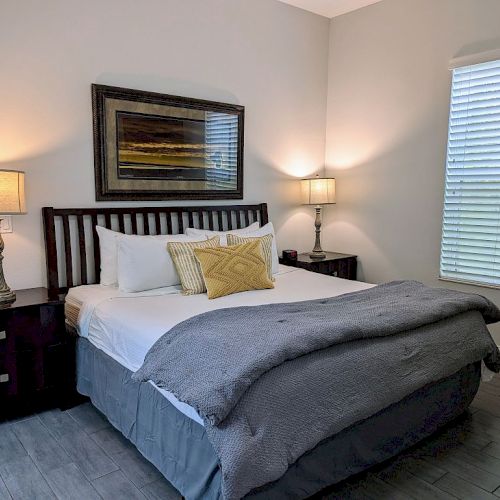 A cozy bedroom with a bed, two bedside tables with lamps, decorative pillows, and a window with blinds.
