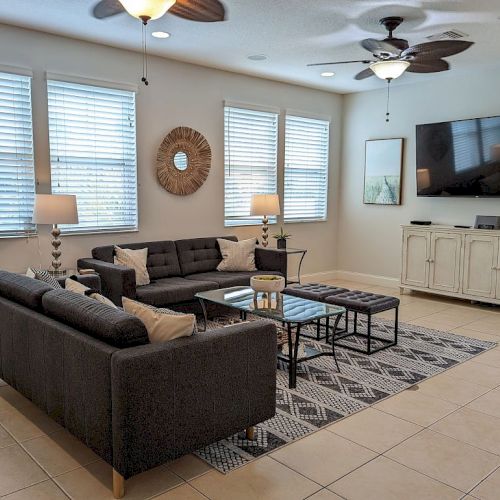 A living room with a dark gray sectional sofa, coffee tables, a large TV, decorative items, and ceiling fans on a tiled floor.