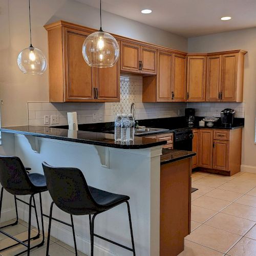 A modern kitchen with wooden cabinets, a countertop with three stools, pendant lights, and windows. There's a coffee maker on the counter.