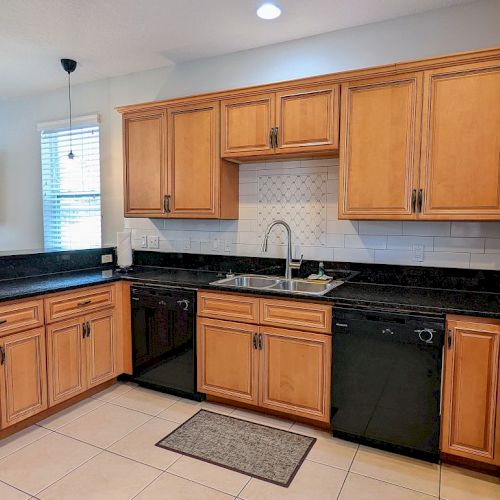 The image shows a kitchen with wooden cabinets, black countertops, white tiles, a sink, and appliances, including a dishwasher and coffee maker.