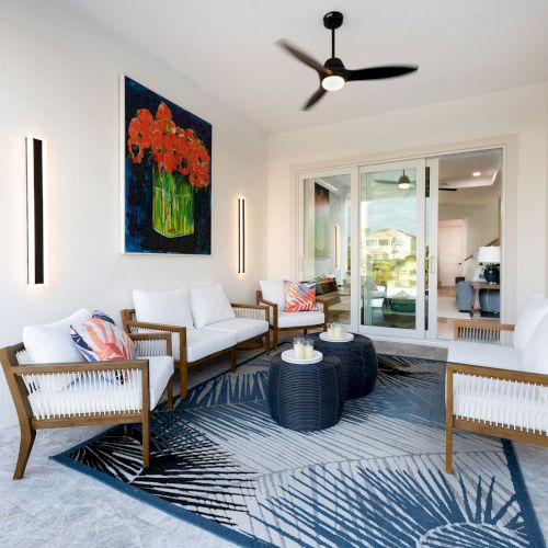 A cozy patio features white seating, a blue rug, modern art, and a ceiling fan, creating a relaxed and stylish outdoor space.