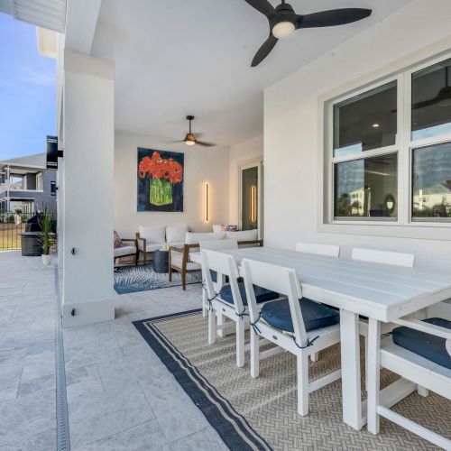 The image shows an outdoor patio with a dining table, chairs, ceiling fan, and decorated walls, by a poolside under a clear sky.