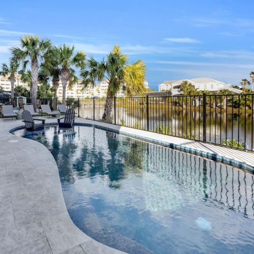 A serene poolside view with lounge chairs, palm trees, and a fence overlooking a waterfront with buildings in the background.