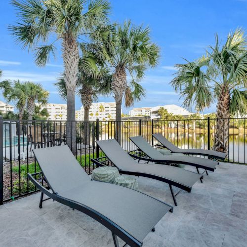 Poolside area with three lounge chairs, palm trees, and a view of buildings in the background under a blue sky.