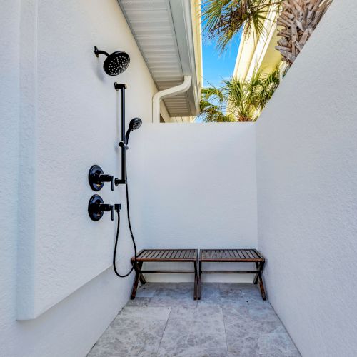 An outdoor shower with black fixtures, white walls, and two wooden stools, surrounded by palm trees under a clear blue sky.