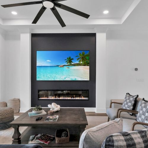 A modern living room with a ceiling fan, TV showing a beach scene, fireplace, cozy seating, and decorative items on a wooden coffee table.