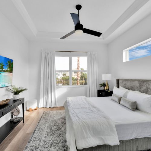 A modern bedroom with a large bed, ceiling fan, flat-screen TV, and a window with a view. Bright and airy atmosphere.