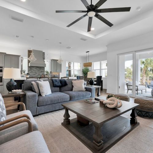 A modern living room with a grey sofa, patterned chairs, a coffee table, and large windows opening to an outdoor view.