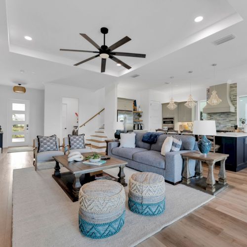A modern living room features a large TV, cozy seating, decorative poufs, and an open kitchen with pendant lights and a center island.