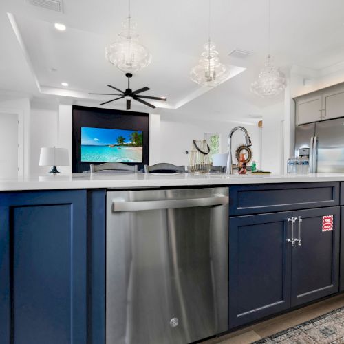 A modern kitchen with a navy island, stainless steel dishwasher, pendant lights, and an open living area featuring a ceiling fan and TV.