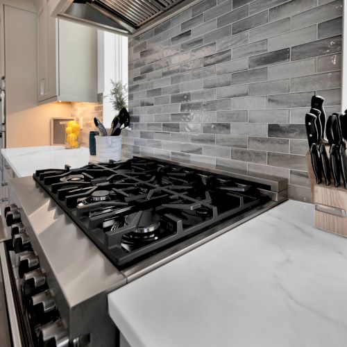 A modern kitchen with a gas stove, marble countertops, gray tiled backsplash, and knife set in a holder on the right.