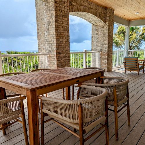 A covered outdoor patio with wooden furniture and brick arches overlooks lush greenery and the ocean, with palm trees in the background.