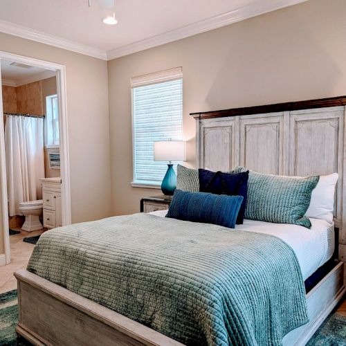 A cozy bedroom with a bed draped in teal accents, two lamps, light wood furniture, and an adjoining bathroom visible through an open door.