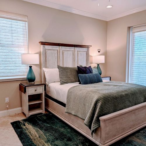 A cozy bedroom with a large bed, two nightstands with lamps, and windows with blinds. The room has light-colored walls and a patterned rug.