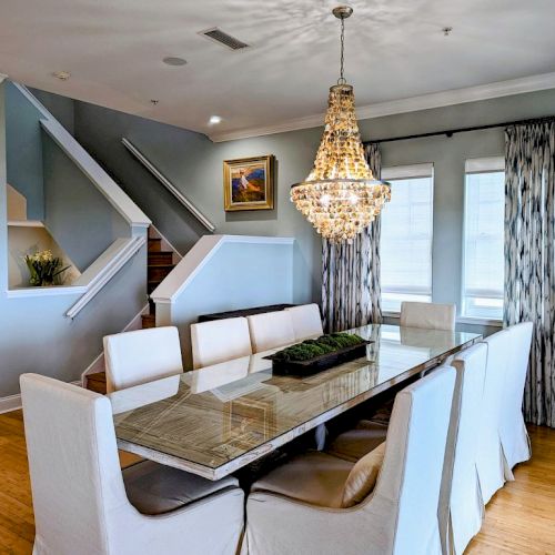 A modern dining room features a large table with white chairs, a chandelier, stairs, and artwork on a gray wall.