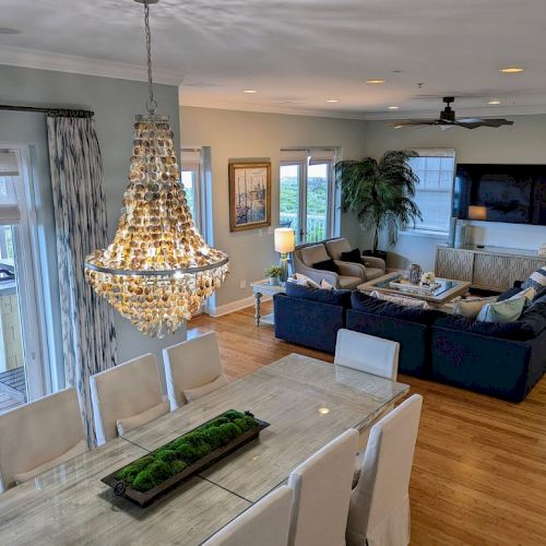 A spacious living and dining area features a large chandelier, a sectional sofa, a TV, and a table with white chairs and a greenery centerpiece.