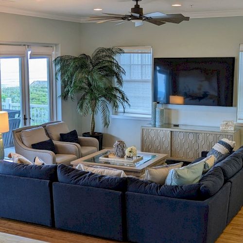 A cozy living room with a large blue sectional sofa, wall-mounted TV, plants, and light decor. The room has wooden flooring.