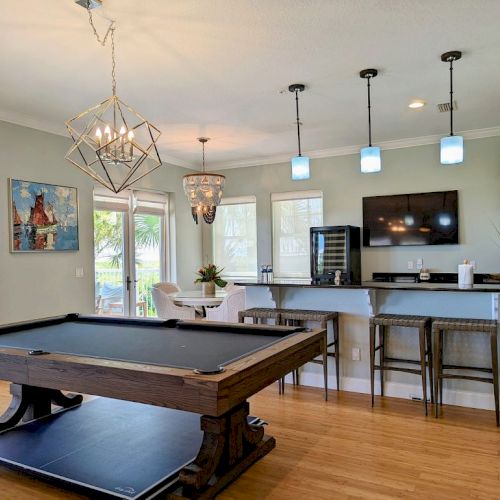 A modern room with a pool table, bar area, high stools, TV, stylish lights, and large windows overlooking a scenic outdoor view.