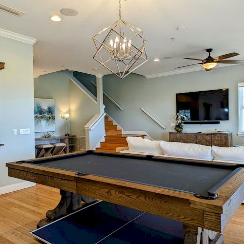 A modern living room with a pool table, a TV, and a couch. There's a staircase, a pendant light, and wall decor.