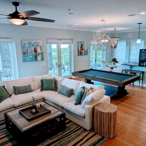 A spacious living room with a sectional sofa, pool table, kitchen bar, and modern decor. Bright windows offer a scenic outdoor view.