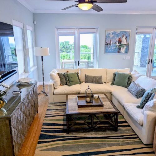 A cozy living room with a white sectional sofa, green pillows, artwork, large windows, and a wooden coffee table on a patterned rug.