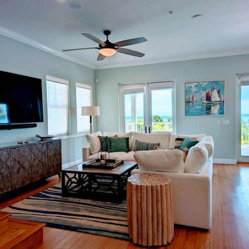 A bright, modern living room with large windows, a sectional sofa, flat-screen TV, and ocean-view balcony doors.