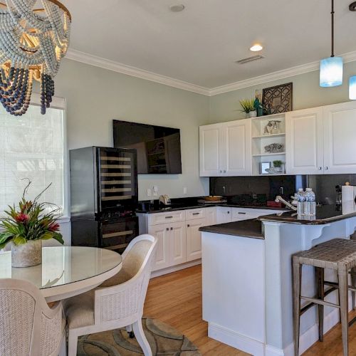 A modern kitchen features white cabinets, black countertops, a dining table with chairs, bar stools, and a decorative chandelier.