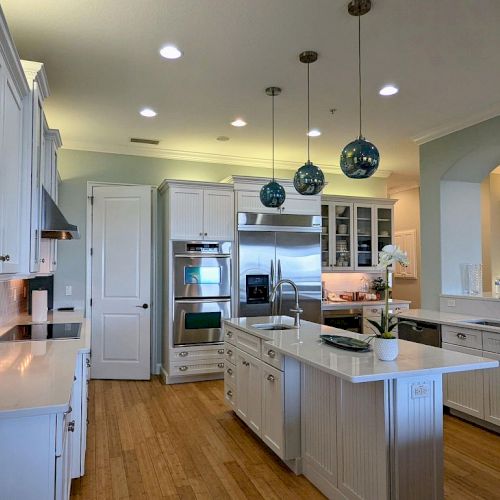 A modern kitchen with white cabinets, an island, pendant lights, stainless steel appliances, and wooden flooring is shown.