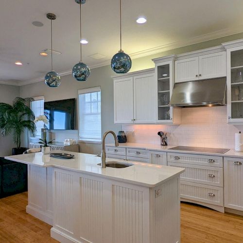 The image shows a modern kitchen with a central island, pendant lights, white cabinets, stainless steel appliances, and an adjacent living area.