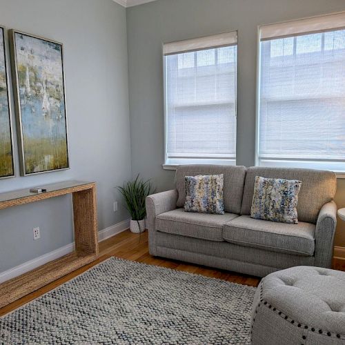 A cozy living room with a gray sofa, abstract art, a rug, side tables, lamp, and natural light.