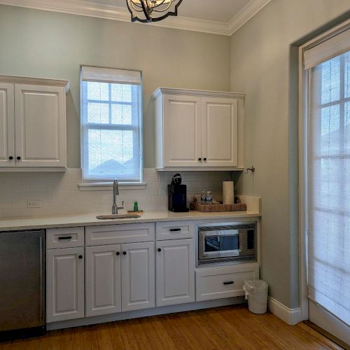 A kitchen with white cabinets, a sink, a microwave, and French doors with sheer curtains, on a wooden floor.
