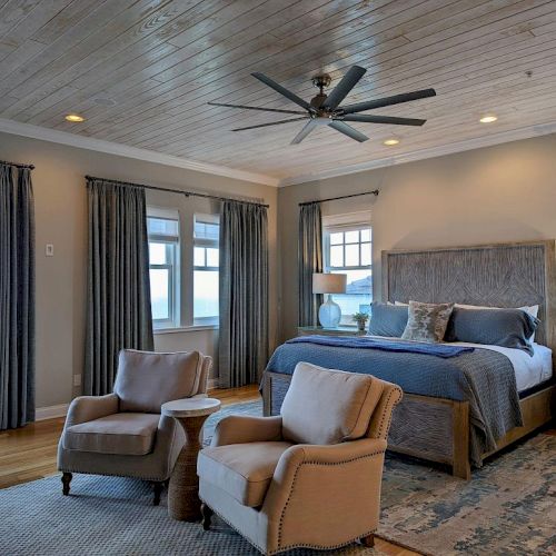 A cozy bedroom with a bed, two armchairs, a ceiling fan, large windows, and bedside tables, featuring soft lighting and a neutral palette.
