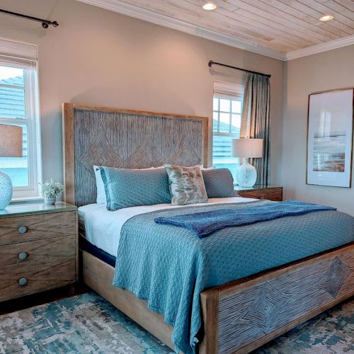 A bedroom with a wooden bed, blue bedding, matching lamps on side tables, large windows, and two wall paintings, all in a cozy setting.