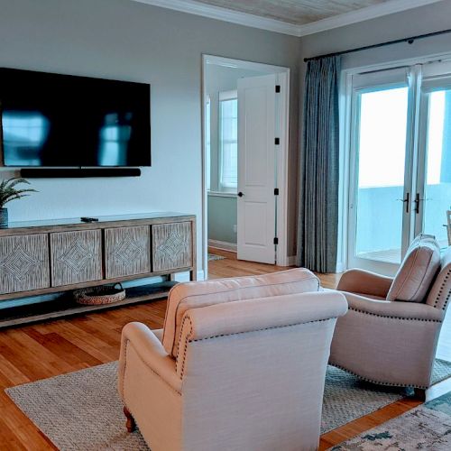The image shows a living room with a wall-mounted TV, two chairs, a console table, and large windows with curtains leading to a balcony.