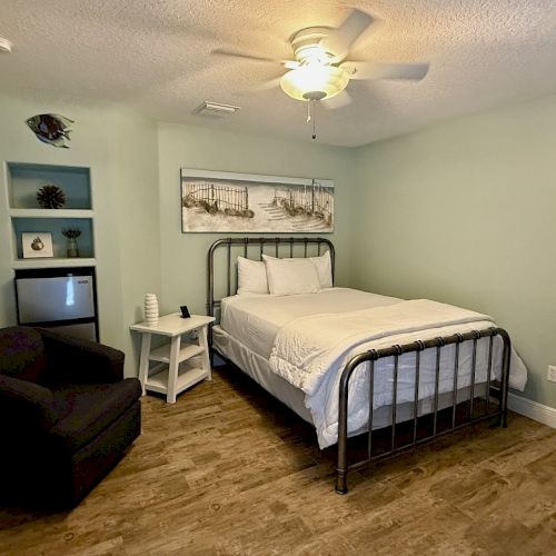 A cozy bedroom with a bed, side table, armchair, wall art, and a built-in shelf with a fridge, against light green walls and hardwood floor.