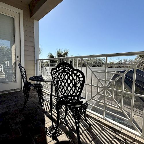 A small balcony with two ornate metal chairs and a table, white railing, and a clear blue sky in the background.