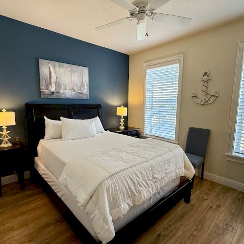 A cozy bedroom features a bed, nautical décor, wooden floor, navy accent wall, two lamps, and large windows with white blinds filtering natural light.