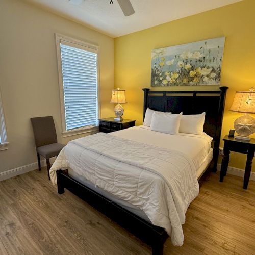 A cozy bedroom features a bed, two bedside tables with lamps, a chair, and a floral painting on a yellow accent wall.