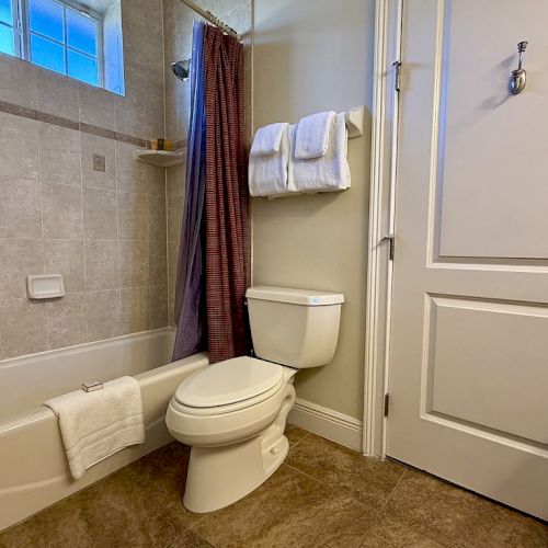 The image shows a bathroom with a tub, toilet, white towels, and a shower curtain. The floor is tiled, and there's a window above the bathtub.