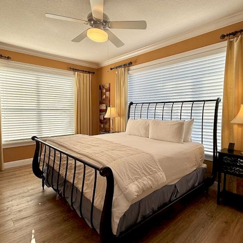 A cozy bedroom with a metal frame bed, white bedding, two lamps, wooden floor, ceiling fan, and large windows with blinds and curtains.