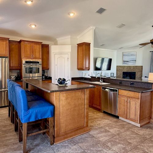 A modern kitchen with wooden cabinets, stainless steel appliances, a central island with blue chairs, and an adjacent living area.