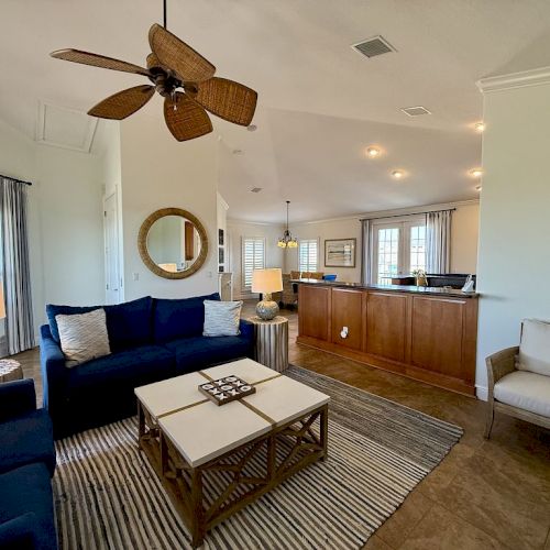 The image shows a cozy living room with a ceiling fan, navy sofa, coffee tables, and a view into a kitchen area with wooden cabinets.