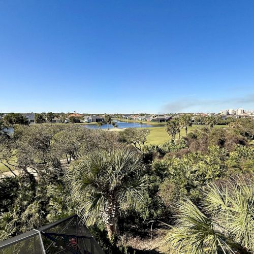 The image shows a scenic view of greenery, trees, and a distant body of water under a clear blue sky.
